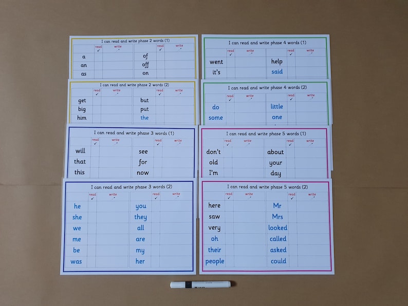 May include: A set of laminated word cards for early reading practice. Each card features a list of words with spaces for reading and writing, organized by phase. The cards are in various colors and include the text "I can read and write phase...words". A black marker is also present.