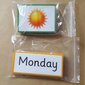 May include: Two sets of flashcards in clear plastic bags. One set features a sun graphic with a green border. The other set has the word "Monday" in black text with an orange border. Educational flashcards.
