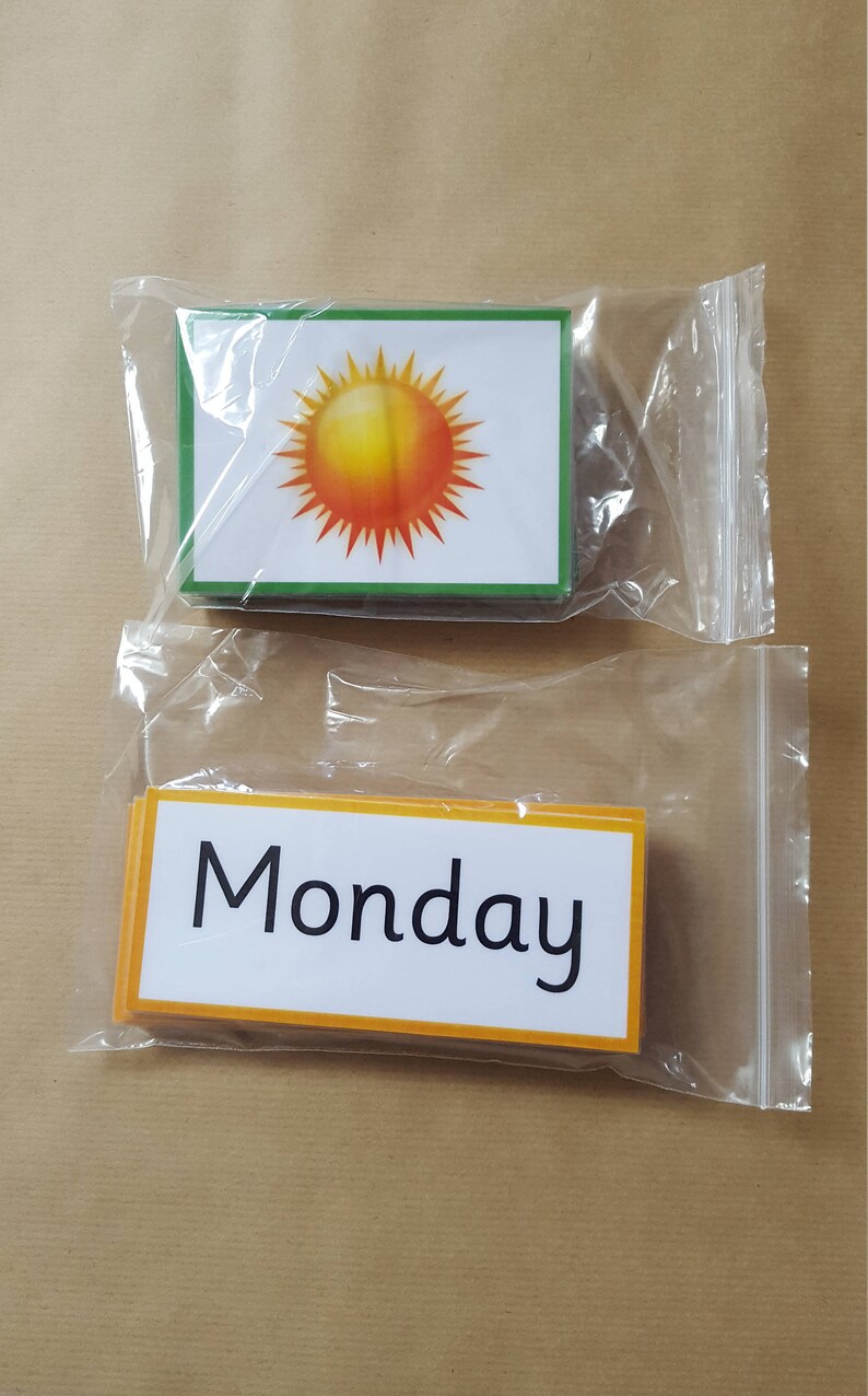 May include: Two sets of flashcards in clear plastic bags. One set features a sun graphic with a green border. The other set has the word "Monday" in black text with an orange border. Educational flashcards.