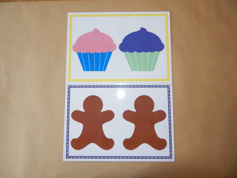 May include: Two cards with illustrations of baked goods. The top card features two cupcakes, one pink and one blue, with blue and green liners. The bottom card shows two brown gingerbread men with white borders and blue trim.