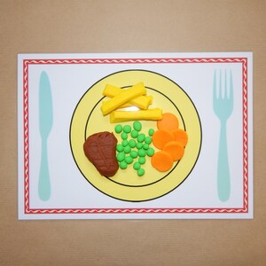 May include: A placemat featuring a yellow plate with a meal of play food: a steak, peas, carrots, and fries. A light blue knife and fork are on either side of the plate. The placemat has a red and white border.