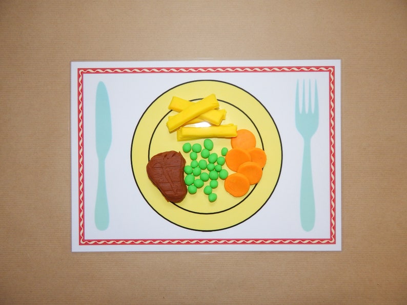 May include: A placemat featuring a yellow plate with a meal of play food: a steak, peas, carrots, and fries. A light blue knife and fork are on either side of the plate. The placemat has a red and white border.