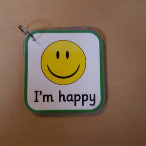 May include: A small, square card with rounded corners and a green border. The card features a yellow smiley face and the text "I'm happy" in black. A metal ring is attached to the top corner.