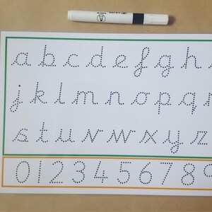 May include: A white paper with the alphabet and numbers in dotted lines for tracing, with a green and orange border. A white and black pen is at the top. Educational material for learning to write.