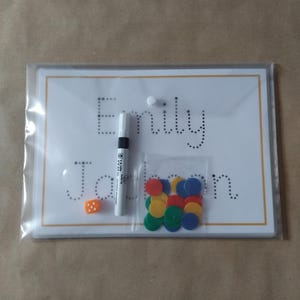 May include: A clear plastic package containing a white board with the name "Emily Jackson" written in dotted letters. A black marker, a small orange die, and a bag of colorful circular counters are also included.