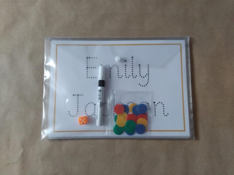 May include: A clear plastic package containing a white board with the name "Emily Jackson" written in dotted letters. A black marker, a small orange die, and a bag of colorful circular counters are also included.