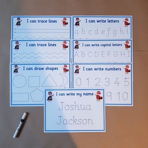 May include: Educational tracing activity set for kids. Includes cards for tracing lines, letters, capital letters, numbers, shapes, and a name. Each card features a superhero graphic and blue borders. A marker is also included.
