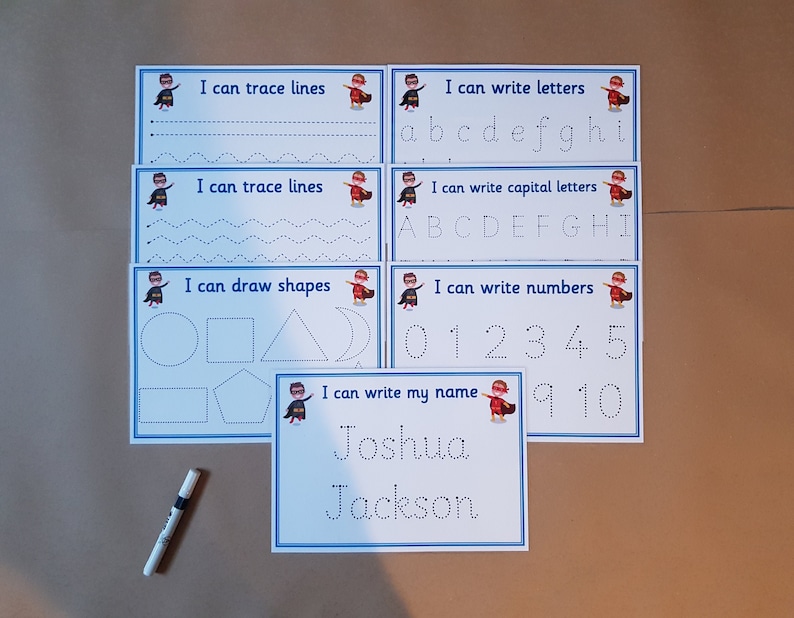 May include: Educational tracing activity set for kids. Includes cards for tracing lines, letters, capital letters, numbers, shapes, and a name. Each card features a superhero graphic and blue borders. A marker is also included.