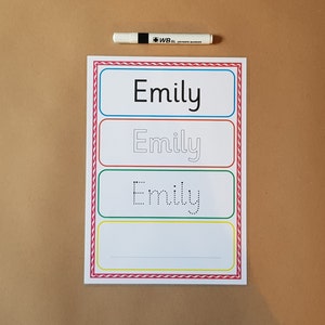 May include: A white learning sheet with the name "Emily" printed in black, with tracing lines for practice. A white dry-erase marker is placed above the sheet. The sheet has a colourful border.