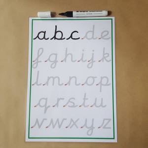 May include: A white sheet of paper with a green border displays lowercase cursive letters a to z. The letters are in grey, with the first four letters in black. A black marker with its cap is placed above the paper.