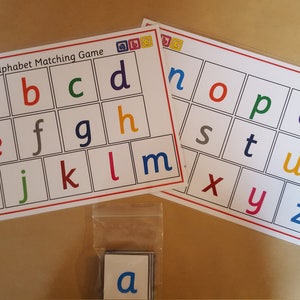 May include: Alphabet matching game featuring two printed sheets with lowercase letters in coloured boxes. A small bag contains matching letter cards. The game is designed for educational purposes.