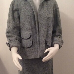 May include: A gray tweed jacket and skirt suit with black trim. The jacket has two flap pockets and the skirt is knee-length.