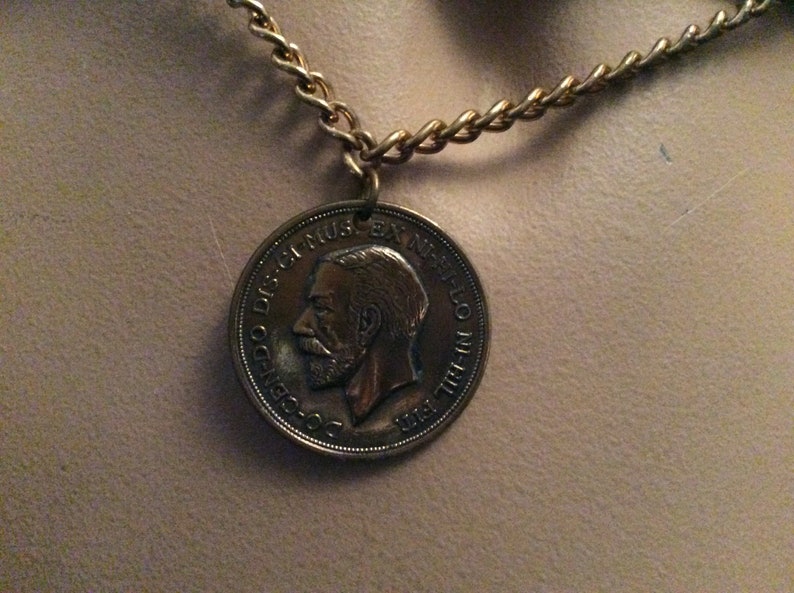 May include: A gold-colored chain necklace with a circular pendant. The pendant is a coin with a profile of a man and text around the edge. The chain is made of small, interlocking links.