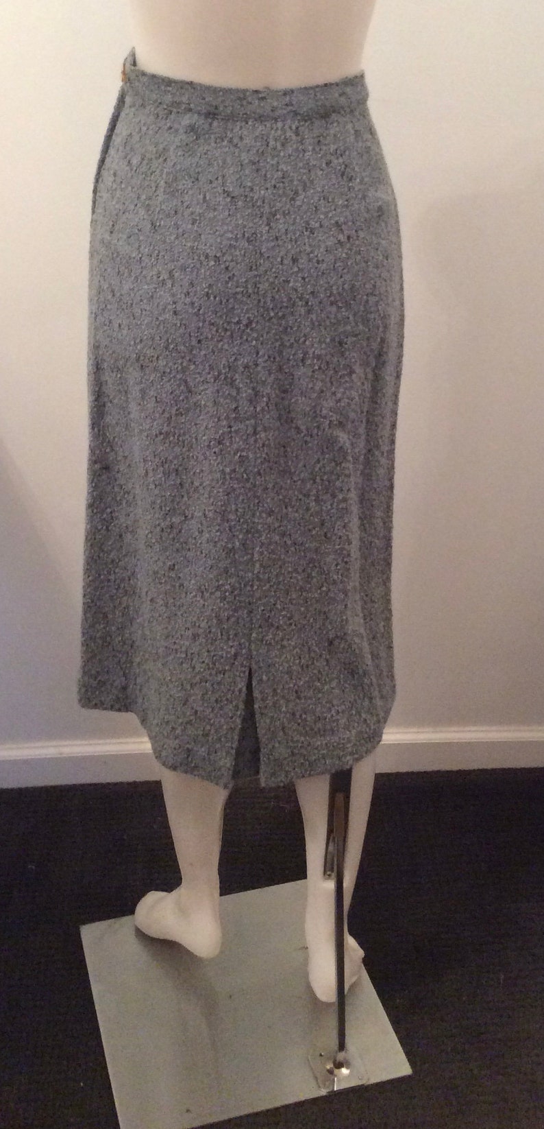 May include: A gray tweed skirt with a back slit. The skirt is on a mannequin and is displayed on a white platform.