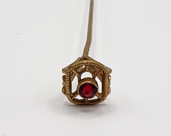 May include: A gold-toned stick pin featuring a square, open-work frame with a beaded border. Centered within the frame is a small, round, deep red gemstone set in a circular bezel. The pin extends upward from the top of the decorative square head.