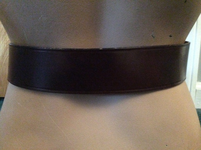 May include: A wide, dark brown belt made of a smooth material. The belt is displayed on a mannequin, showcasing its shape and design. The belt's color is a deep, rich brown, suitable for various outfits.