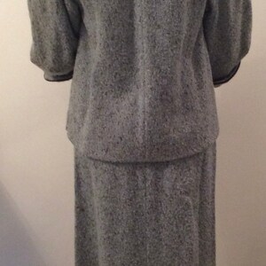 May include: A gray tweed skirt suit with a black trim. The jacket has a collar and three-quarter sleeves. The skirt is knee-length.