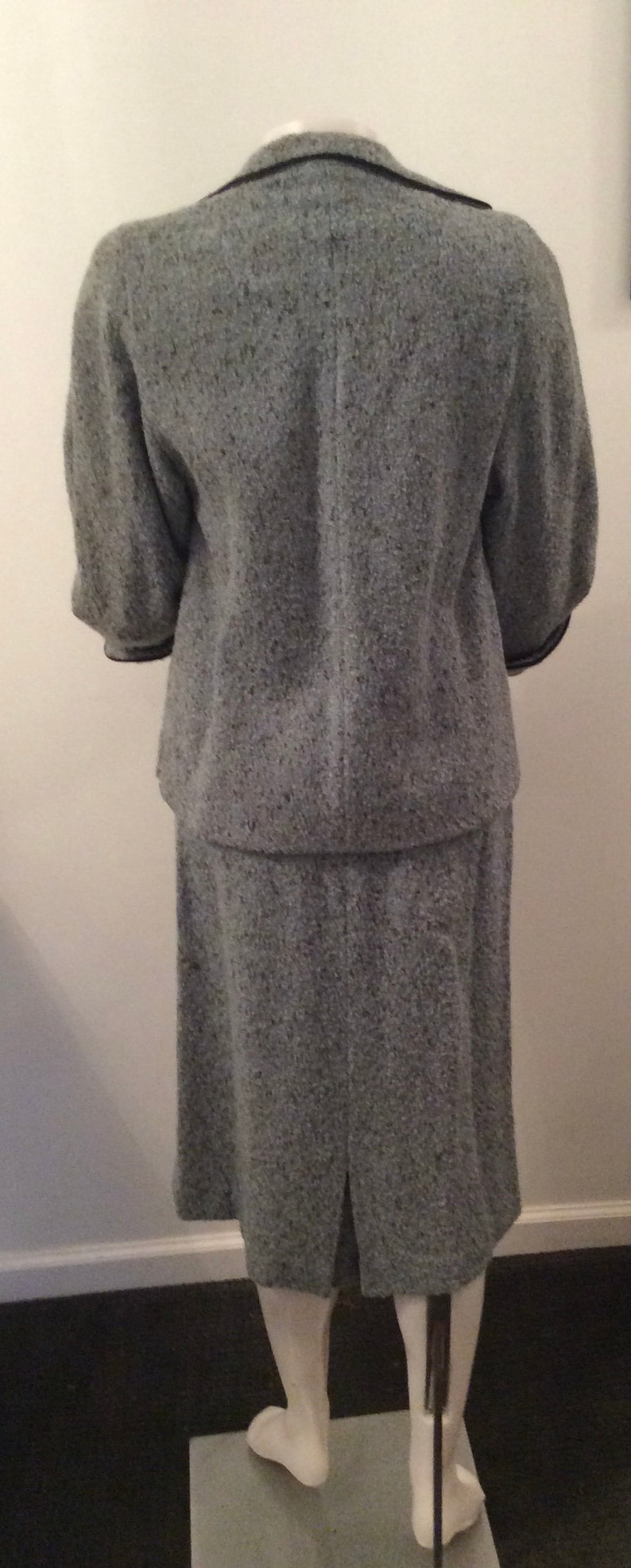 May include: A gray tweed skirt suit with a black trim. The jacket has a collar and three-quarter sleeves. The skirt is knee-length.