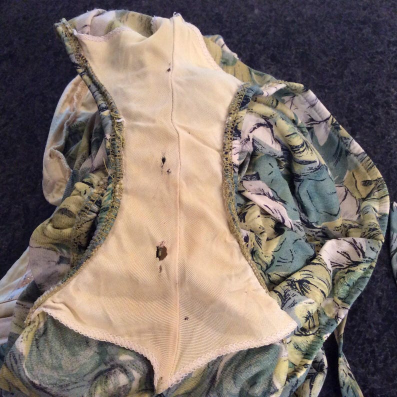 May include: A close-up of the inside of a green and yellow floral print dress. The dress has a yellow lining and a seam down the center back. There are a few small brown stains on the lining.