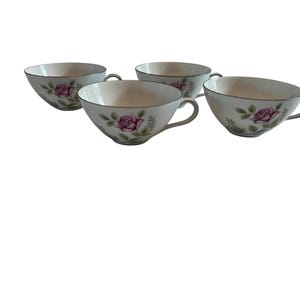May include: Four white teacups with a floral rose design. Each cup has a silver rim and handle, and a pink rose with green leaves is painted on the side. The cups are arranged on a white background.