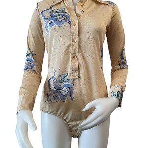 May include: A tan bodysuit with long sleeves and a collared neckline. The bodysuit features a button-down front and is adorned with a blue and white dragon design on the front and sleeves. The fabric has a speckled pattern.