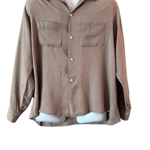 May include: A tan-colored long-sleeve shirt with two front pockets and a button-down closure. The shirt is displayed on a white mannequin and has a classic collar. The fabric appears to have a subtle sheen.