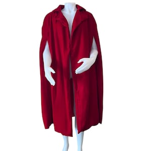 May include: A vibrant red cape with a collar and open front, displayed on a mannequin. The cape features arm slits and a flowing design, perfect for costumes or fashion. The fabric appears to be a soft, velvety material.