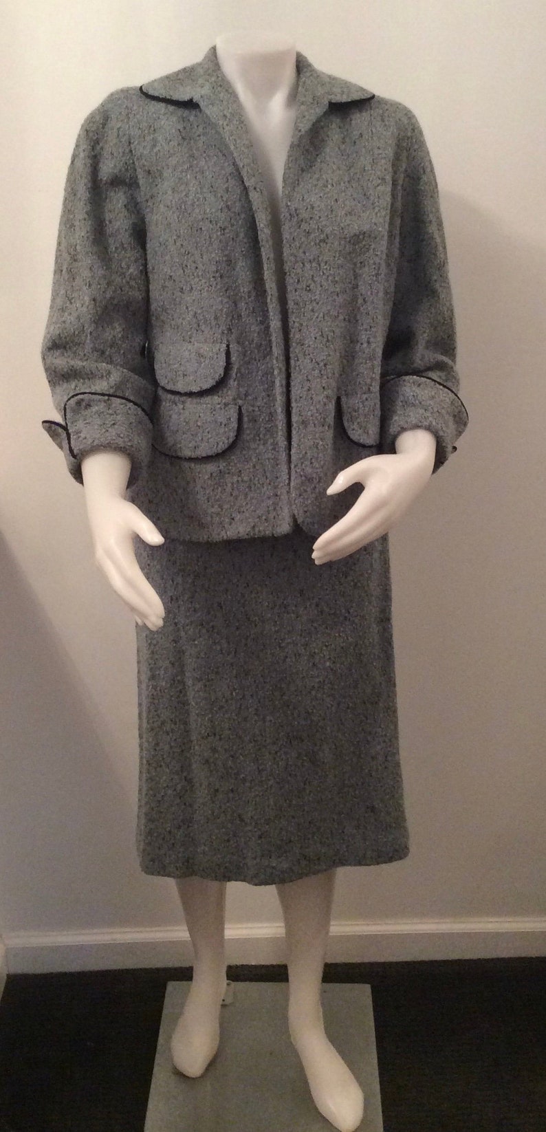 May include: A gray tweed jacket and skirt suit with black trim. The jacket has two flap pockets and the skirt is knee-length.