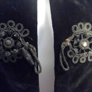May include: A black velvet jacket with two decorative black fabric rosettes with black beads and a gold button in the center.