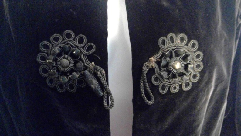 May include: A black velvet jacket with two decorative black fabric rosettes with black beads and a gold button in the center.