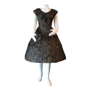 May include: A knee-length dress featuring a dark, intricate circular pattern over a light tan base. The garment has short cap sleeves, a fitted bodice with a central lace-up detail at the neckline, and a full, flared skirt silhouette.