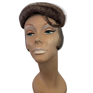 May include: A brown tweed hat with a curved feather accent. The hat is displayed on a mannequin head with painted features, including blue eyes and light pink lips. The hat has a rounded crown.