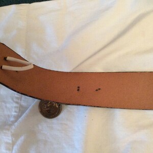 May include: A brown leather belt with a textured, dark blue trim. The belt has two white loops near the buckle end. A gold coin rests near the belt. The belt is on a white surface.