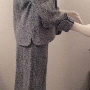 May include: A gray tweed jacket and skirt suit with black trim. The jacket has a collar and long sleeves. The skirt is knee-length.