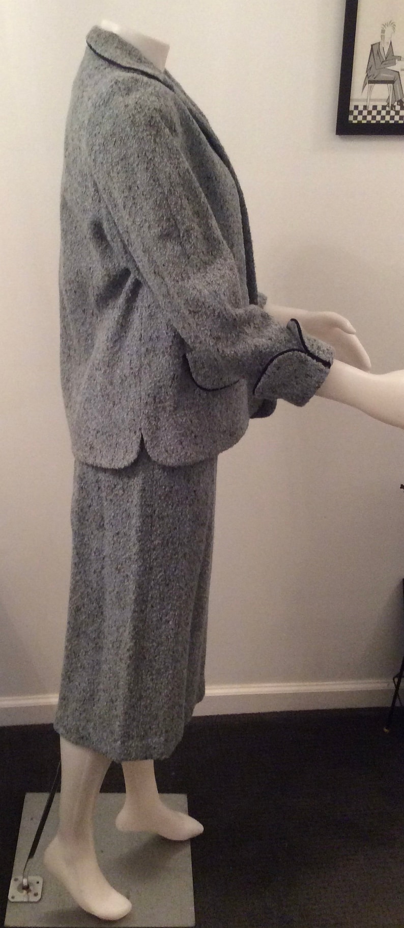 May include: A gray tweed jacket and skirt suit with black trim. The jacket has a collar and long sleeves. The skirt is knee-length.