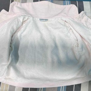 K&ouml;nnte beinhalten: Hellrosa Jacke mit wei&szlig;em Innenfutter. Die Jacke hat einen Kragen und Kn&ouml;pfe vorne. Das Innere besteht aus weichem, pl&uuml;schigem Material. Die Jacke ist offen und zeigt das Innere.