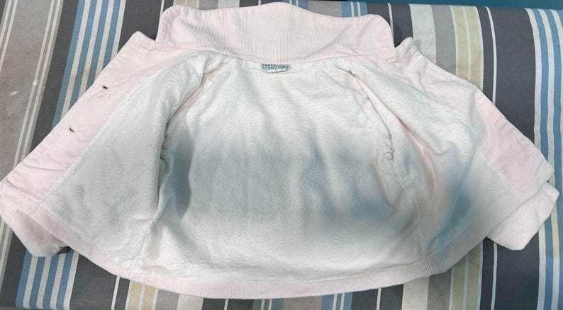 K&ouml;nnte beinhalten: Hellrosa Jacke mit wei&szlig;em Innenfutter. Die Jacke hat einen Kragen und Kn&ouml;pfe vorne. Das Innere besteht aus weichem, pl&uuml;schigem Material. Die Jacke ist offen und zeigt das Innere.
