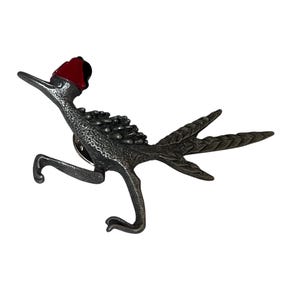May include: A metallic silver-toned bird-shaped pin featuring a textured body, long tail feathers, and a distinct red crest on its head. The pin is depicted in a running pose with one leg raised and the other extended downward, showcasing a detailed finish.