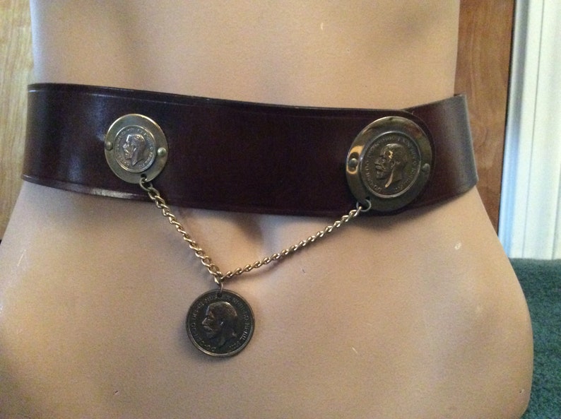 May include: A brown leather belt with three decorative coin medallions. The belt has a wide band and is fastened with two circular medallions. A gold chain connects the medallions to a third, hanging coin.