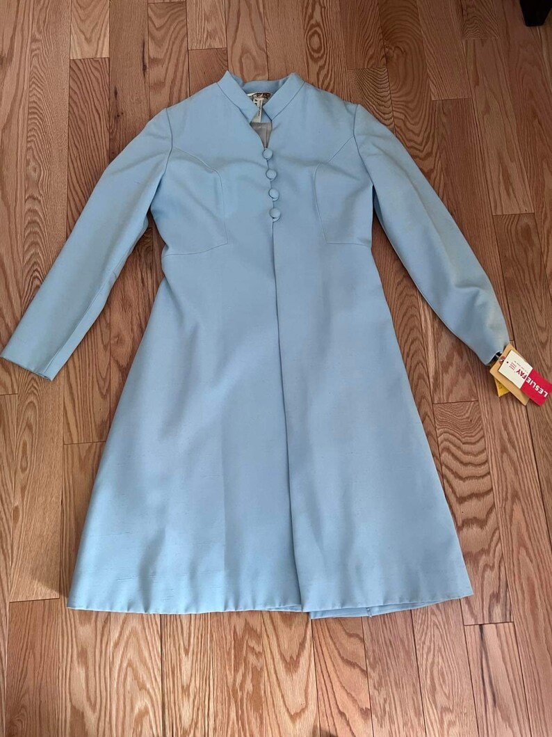 May include: A light blue long-sleeved coat with a mandarin collar and button closure. The coat has a fitted bodice and a flared skirt. The coat is laying flat on a wooden floor.