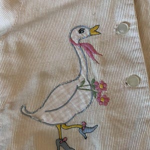 K&ouml;nnte beinhalten: Nahaufnahme eines hellrosa Cordkleidungsst&uuml;cks mit einem skurrilen, gestickten G&auml;nsemotiv. Die Gans tr&auml;gt einen rosa Schal und tr&auml;gt Blumen. Das Kleidungsst&uuml;ck hat wei&szlig;e Kn&ouml;pfe und ein vertikales Streifenmuster.