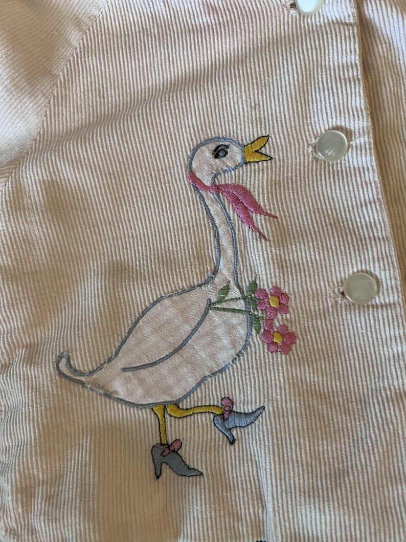 K&ouml;nnte beinhalten: Nahaufnahme eines hellrosa Cordkleidungsst&uuml;cks mit einem skurrilen, gestickten G&auml;nsemotiv. Die Gans tr&auml;gt einen rosa Schal und tr&auml;gt Blumen. Das Kleidungsst&uuml;ck hat wei&szlig;e Kn&ouml;pfe und ein vertikales Streifenmuster.