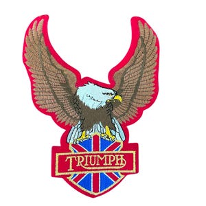 May include: An embroidered patch featuring a bald eagle with brown wings and a white head, perched above a shield displaying the Union Jack flag. The word TRIUMPH is stitched in white letters on a red rectangular banner across the shield.