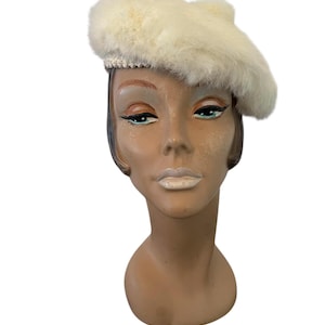 May include: A cream-colored beret made of fluffy material. The hat features a decorative band around the base. The beret is displayed on a mannequin head with painted features, including green eyes and pale lips. The background is white.