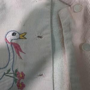 K&ouml;nnte beinhalten: Nahaufnahme einer hellrosa Cordjacke mit wei&szlig;em Futter. Die Jacke hat ein gesticktes G&auml;nsemotiv mit rosa Schal und Blumen. Sie hat wei&szlig;e Kn&ouml;pfe und einen hellrosa Kragen.