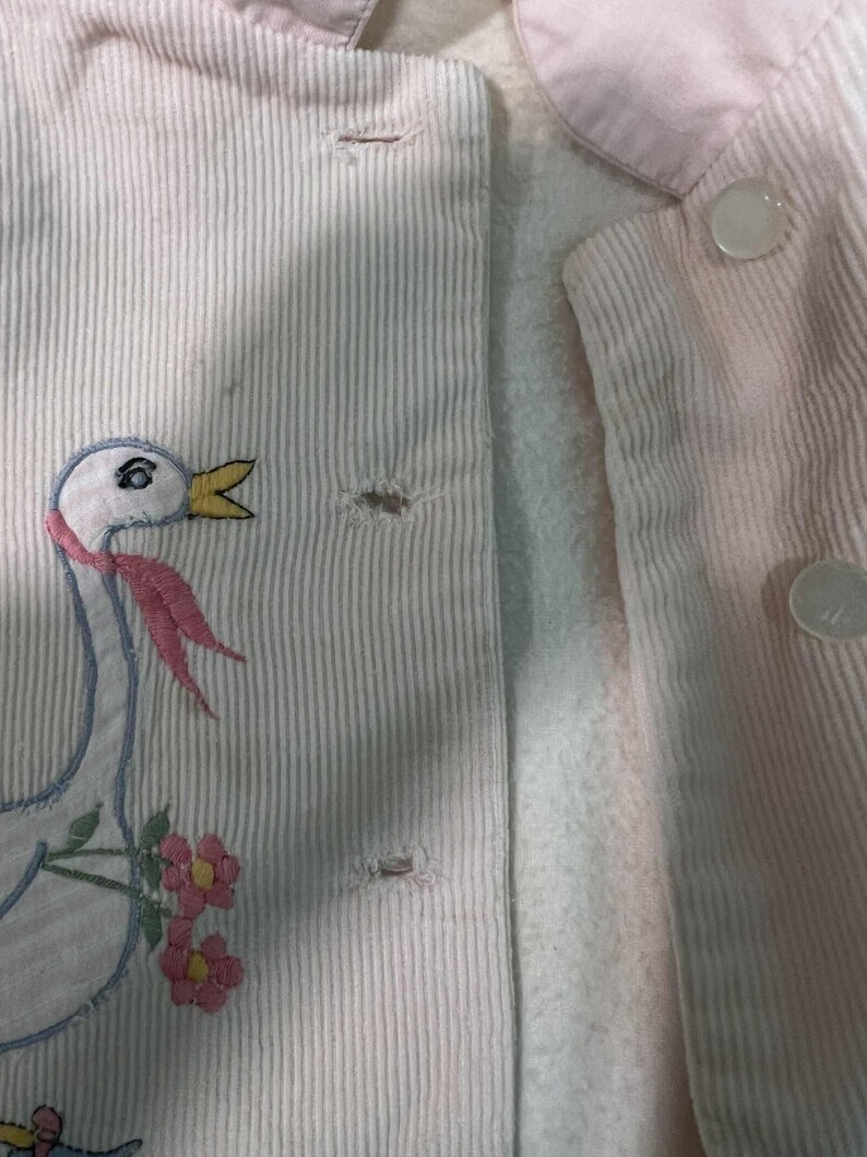 K&ouml;nnte beinhalten: Nahaufnahme einer hellrosa Cordjacke mit wei&szlig;em Futter. Die Jacke hat ein gesticktes G&auml;nsemotiv mit rosa Schal und Blumen. Sie hat wei&szlig;e Kn&ouml;pfe und einen hellrosa Kragen.