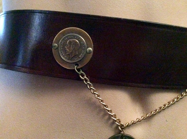 May include: A dark brown leather belt with a circular bronze medallion featuring a profile portrait and text. A gold-colored chain hangs from the medallion. The belt is wide and has a classic design.