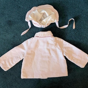 K&ouml;nnte beinhalten: Hellrosa Baby-Jacke und passendes M&uuml;tzenset. Die Jacke hat lange &Auml;rmel und einen Kragen. Die M&uuml;tze hat Bindeb&auml;nder und ein Satinfutter. Das Set ist aus einem weichen, m&ouml;glicherweise Cord-Material.