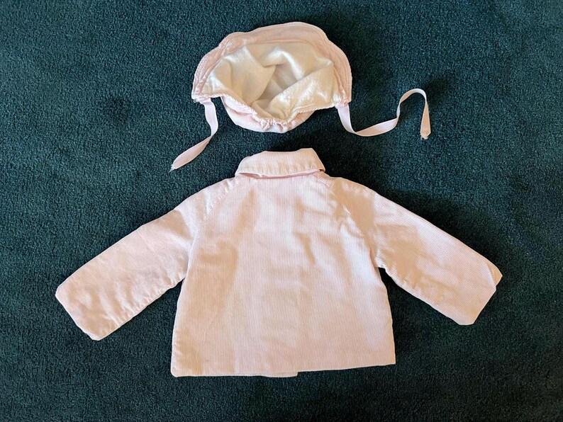 K&ouml;nnte beinhalten: Hellrosa Baby-Jacke und passendes M&uuml;tzenset. Die Jacke hat lange &Auml;rmel und einen Kragen. Die M&uuml;tze hat Bindeb&auml;nder und ein Satinfutter. Das Set ist aus einem weichen, m&ouml;glicherweise Cord-Material.