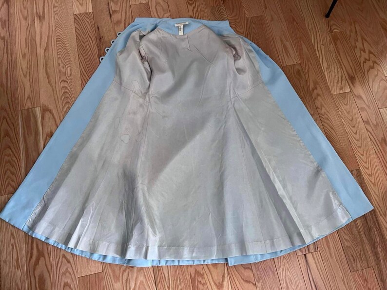 May include: A light blue coat with a white lining. The coat is open and the lining is visible. The coat has a button closure and a long, flowing skirt.